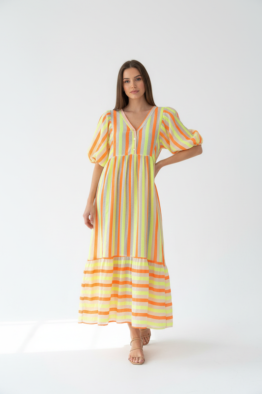 Model in neon gestreepte maxi-jurk You Passion met geel, oranje en grijs, gestileerd met nude sandalen